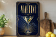 A framed poster of a martini glass with an olive on the rim, placed on a wooden surface next to a wooden cutting board and a potted plant.
