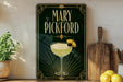 A framed poster of a cocktail called "Mary Pickford" is displayed on a wall, with a wooden cutting board and a potted plant to the side.