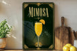 A framed poster of a mimosa cocktail is displayed on a wall, with a wooden cutting board and a potted plant to the side.