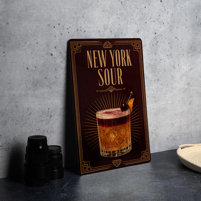 A decorative sign for "New York Sour" cocktail, featuring an illustration of a cocktail glass with a cherry on top, placed on a gray countertop next to some black camera lenses.