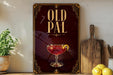 a poster or sign with the text "OLD PAL" and an illustration of a glass of red liquid, possibly a cocktail or wine, with a lemon slice. The poster is displayed on a wall, and there are some kitchen utensils and a potted plant visible in the background.