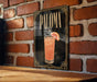 A framed poster of a cocktail called "Paloma" is displayed on a brick wall.