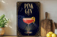A framed poster of a pink gin cocktail is displayed on a wall, with a wooden cutting board and a potted plant to the side.