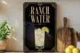 A decorative sign for Ranch Water, featuring a glass of water with a lime slice, is displayed on a wooden shelf next to a cutting board and a potted plant.