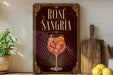 a decorative sign for "Rose Sangria" with a glass illustration of a rose-infused drink, placed on a wooden surface next to a potted plant and a cutting board.