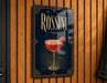 A framed poster or sign featuring a red cocktail with a strawberry garnish, hanging on a wooden wall.