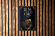 A poster with the text "RUM AND COKE" and an illustration of a glass of rum and coke on a wooden wall.