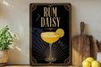 a framed poster with an illustration of a cocktail glass and a lemon slice, along with the text "RUM DAISY" on a black background with gold accents.