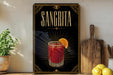 A framed poster of a Sangrita cocktail is displayed on a wall, with a wooden cutting board and a potted plant to the side.