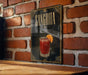 A framed poster of a Sangrita cocktail hangs on a brick wall, featuring an illustration of a glass filled with the drink and an orange slice.