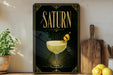 A framed poster of a cocktail called "Saturn" is displayed on a wall, with a lemon and a wooden cutting board visible to the right.