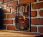 A poster of a drink called "Sazerac" is displayed on a brick wall, with a glass of the drink and a lemon peel in the center of the poster.