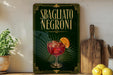 a poster or sign with the text "SBAGLIATO NEGRONI" and an illustration of a glass of Negroni, a type of Italian cocktail. The poster is displayed on a wall, and there are some kitchen utensils, such as a knife and a wooden cutting board, as well as some fruits, such as lemons, in the foreground.