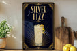 A framed advertisement for a "Silver Fizz" cocktail, featuring a glass of the drink with a lemon twist and a decorative border.