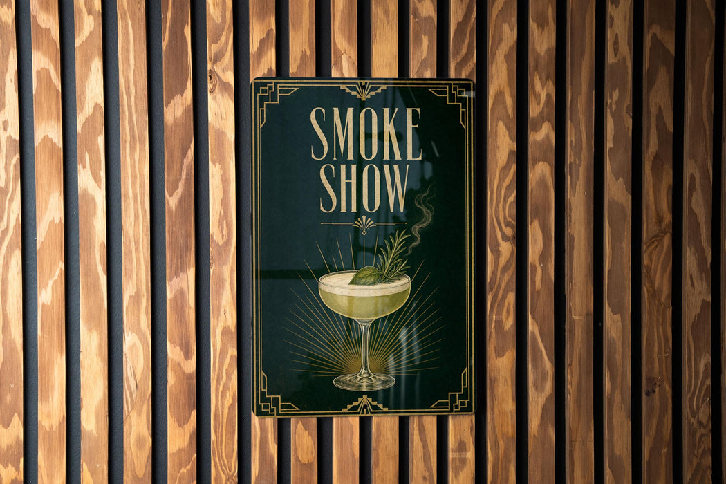 A poster with the text "SMOKE SHOW" and an illustration of a cocktail glass with a plant in it, displayed on a wooden wall.
