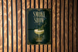 A poster with the text "SMOKE SHOW" and an illustration of a cocktail glass with a plant in it, displayed on a wooden wall.
