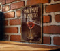 A framed poster of a smoked maple Manhattan cocktail is displayed on a brick wall.