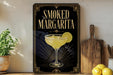 A framed poster advertising a "Smoked Margarita" is displayed on a wall, with a potted plant and wooden cutting board visible in the foreground.