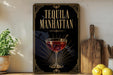 A framed poster with the text "TEQUILA MANHATTAN" and an illustration of a tequila drink, placed on a wooden surface next to a cutting board and a potted plant.