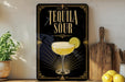 A decorative sign with the text "TEQUILA SOUR" and an illustration of a tequila sour cocktail, along with a lemon slice, is displayed on a wooden shelf.