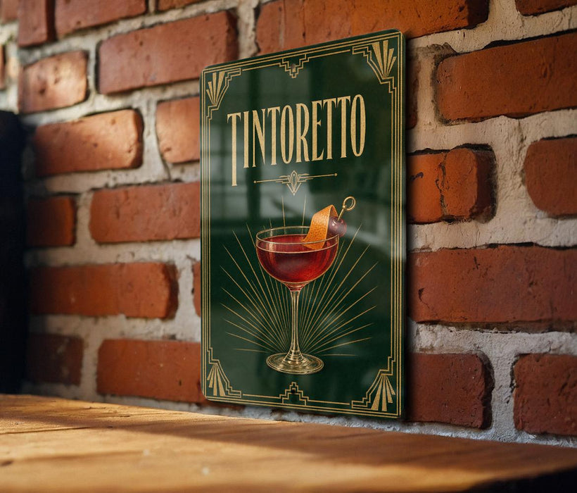 A framed poster of a Tintoretto cocktail hangs on a brick wall, with a wooden table below it.