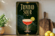 A framed poster of a cocktail called "Trinidad Sour" is displayed on a wall, with a wooden cutting board and a potted plant to the side.