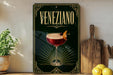 a decorative sign with the word "Veneziano" prominently displayed, along with an illustration of a cocktail glass and garnish. The sign is placed on a wooden surface, with a potted plant, a cutting board, and some lemons nearby.