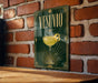 A framed poster of a cocktail glass with a yellow drink inside, hanging on a brick wall.