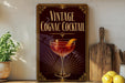 A vintage-style sign advertising a cognac cocktail, featuring an illustration of a glass filled with a red-colored drink and an orange peel.