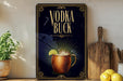 a decorative sign with the text "VODKA BUCK" and an illustration of a copper mug with a lime slice. The sign is displayed on a shelf, with a potted plant, a wooden cutting board, and some lemons nearby.