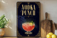 A decorative sign for a vodka punch, featuring a glass of the drink and garnishes, is displayed on a shelf next to a wooden cutting board and a potted plant.