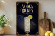 a decorative sign for vodka rickey, with a glass of the drink and a lemon slice, placed on a wooden surface next to a cutting board and some lemons.