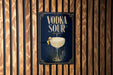 A sign with the text "VODKA SOUR" and an illustration of a cocktail glass with a lemon twist, mounted on a wooden wall.
