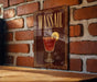 A framed poster of a red cocktail with a lemon slice, displayed on a brick wall.