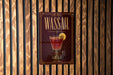 A metallic sign with the word "WASSail" on it, featuring an illustration of a cocktail glass with a lemon slice.