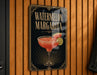 A sign for a "Watermelon Margarita" cocktail is displayed on a wooden wall.