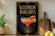 A framed poster of a watermelon margarita is displayed on a wall, with a wooden cutting board and a potted plant to the side.