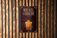 A sign for a whiskey cobbler cocktail is displayed on a wooden wall.