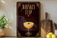 A decorative sign with the text "WHISKEY FLIP" and an illustration of a cocktail glass with a lemon slice, placed on a wooden surface next to a potted plant and a cutting board.