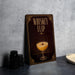 a decorative sign with a whiskey flip cocktail illustration, placed on a countertop next to some black camera lenses.