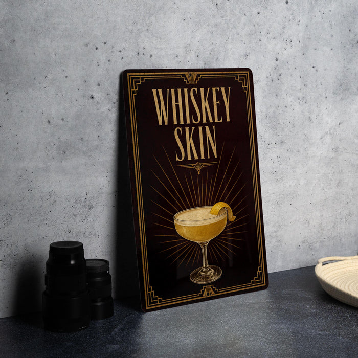 A decorative sign with the text "WHISKEY SKIN" and an illustration of a cocktail glass with a lemon twist, placed on a gray countertop next to some black camera lenses.