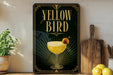 A framed poster with the text "YELLOW BIRD" and an illustration of a cocktail glass with a lemon slice on top, placed on a wooden shelf next to a wooden cutting board and a potted plant.