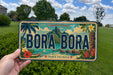 A hand holding a license plate with the text "BORA BORA" and a tropical island landscape in the background.