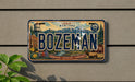 A vintage Montana license plate with the word "BOZEMAN" prominently displayed, featuring various Montana-themed illustrations such as a water tower, mountains, and a moose.