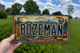 A person is holding a Montana license plate with the word "BOZEMAN" prominently displayed.