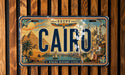 A vintage license plate with the word "CAIRO" prominently displayed, featuring an illustration of a cityscape with landmarks such as the pyramids of Giza and the sphinx.
