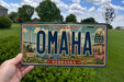A person is holding a license plate from Nebraska that says "OMAHA NEBRASKA" with a scenic illustration of the state's landmarks and natural beauty.