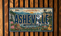 The image is a vintage license plate from North Carolina that features the city of Asheville, with a scenic mountain landscape and a castle in the background.
