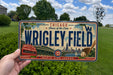 A person is holding a Chicago Cubs-themed license plate that says "WRIGLEY FIELD" and features an illustration of the Wrigley Field stadium and the Chicago Cubs logo.
