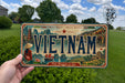 A vintage Vietnamese license plate is being held up against a backdrop of trees and a blue sky with clouds.